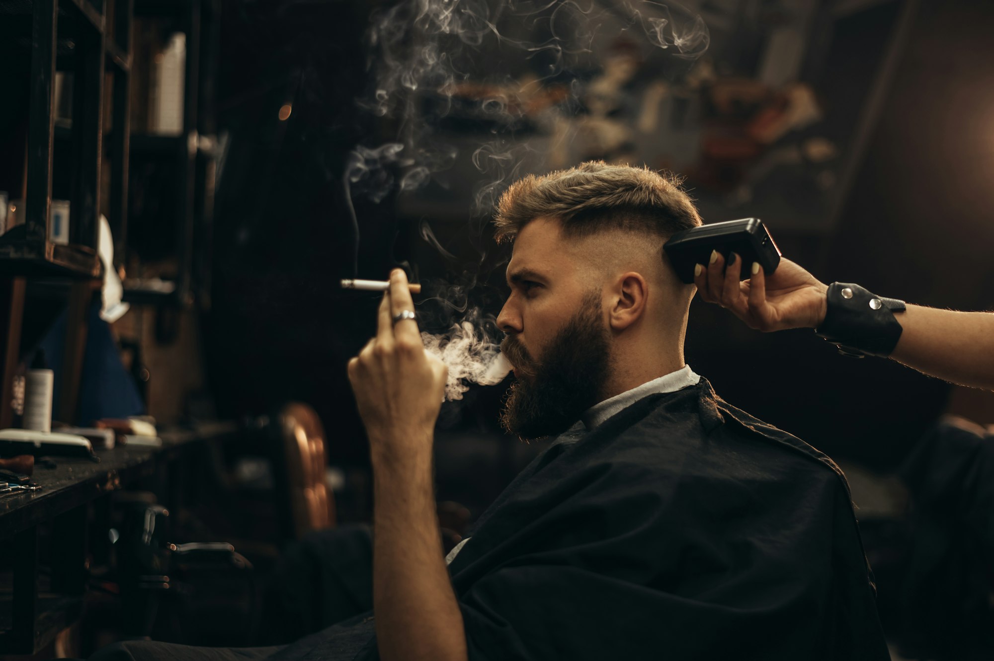 Young bearded man getting haircut by hairdresser and smoking a cigarette